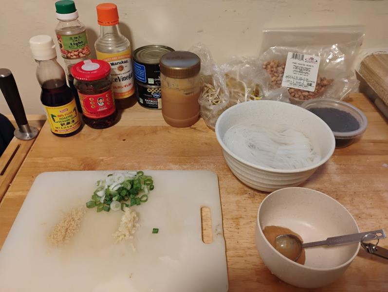 A kitchen prep counter displays the mise en place: Bottles of light soy sauce, rice vinegar, chili crisp, roasted sesame oil; peanut butter; cans of water chestnuts and bamboo shoots; bags of mung and soybean sprouts, honey roasted peanuts; a container of black sesame seeds. In the foreground are a bowl of about 2 (dry) oz soaking mei fun noodles, a cutting board with grated ginger, pressed garlic, and a sliced scallion, and a sauce bowl with the first tablespoon of peanut butter added.
