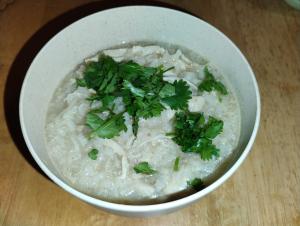 A small bowl holding a stew of rice and shredded chicken, garnished with cilantro. The broth has a lovely shimmer and the rice is cooked until nearly falling apart.